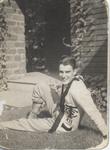 A young man relaxing on a lawn, wearing what appears to be a cardigan from a school uniform. The cardigan displays the letters "T C S".